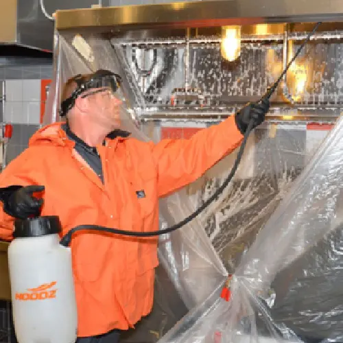 A man in an orange jacket spraying disinfectant on a commercial kitchen hood.